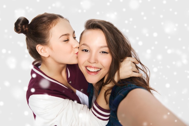 winter, christmas, people, teens and friendship concept - happy smiling pretty teenage girls or friends taking selfie and kissing over gray background and snow