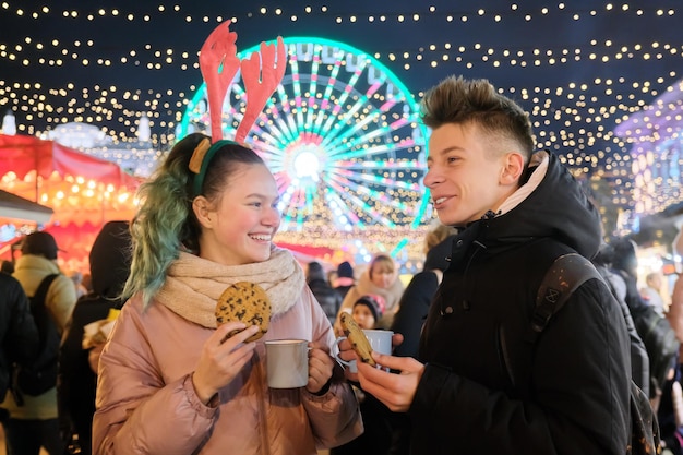 Winter Christmas portrait of a happy teenagers boy and girl at holiday market, laughing teens with cups of hot tea and chocolate gingerbread. Lights, bright garlands of evening city, ferris wheel