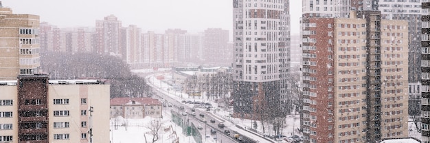 雪が降る冬の街の風景。高さからの町の眺め。雪片のある通りの吹雪。住宅と車を運転する道路。バナー