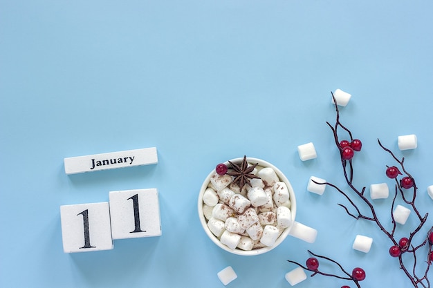 Composizione invernale. cubi calendario in legno bianco. dati 11 gennaio. tazza di cacao, marshmallow e ramo decorativo con bacche rosse su sfondo blu vista dall'alto spazio di copia piatta