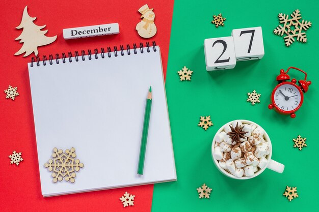 Winter composition. Wooden calendar December 27th Cup of cocoa with marshmallow, empty open notepad with pencil, snowflake, alarm clock on red and green background. Top view Flat lay Mockup