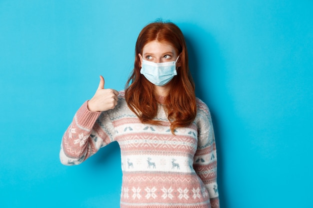 Inverno, coronavirus e concetto di distanza sociale. bella ragazza dai capelli rossi con maschera facciale e maglione, guardando la promozione nell'angolo in alto a sinistra e mostrando il pollice in su, lodando il buon prodotto.