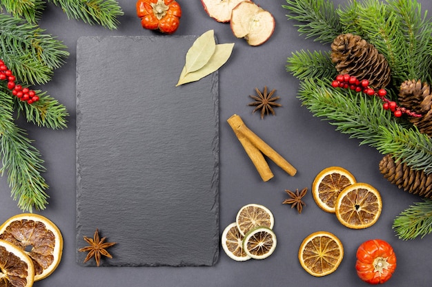 겨울 말린 과일과 요리 향신료 배경에는 크리스마스 나무 가지와 소나무 콘이 있는 어두운 배경에 검정색 슬레이트 칠판이 있습니다. 평면도. 공간을 복사합니다.