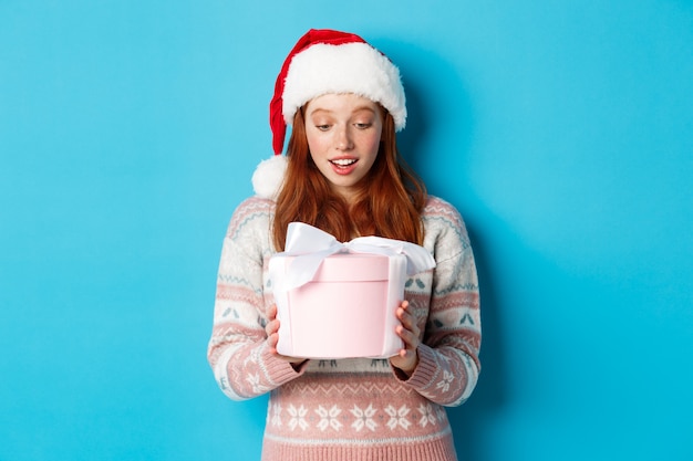 Winter en kerstavond concept. Aangetast en gevleid roodharige meisje kijken naar doos met kerst cadeau, glimlachend verbaasd, staande in kerstmuts tegen blauwe achtergrond.