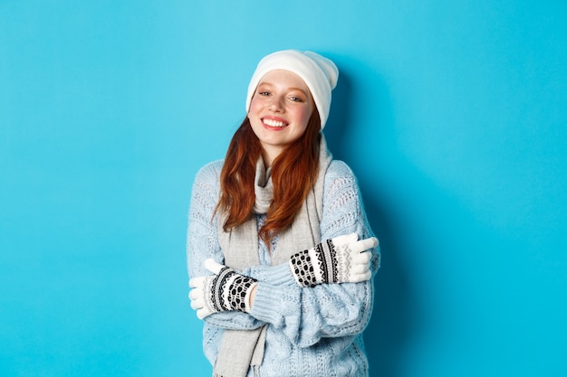 Winter en vakantie concept. Glimlachend roodharig meisje in muts, handschoenen en trui die warm wordt nadat ze naar buiten is gegaan, staande over een blauwe achtergrond.
