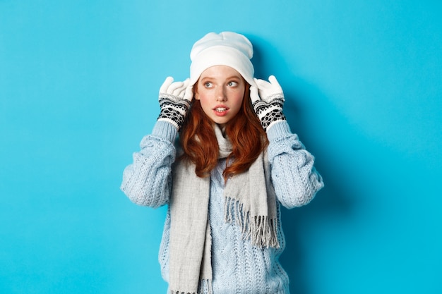 Winter en vakantie concept. Leuk roodharig meisje dat naar buiten gaat, muts en handschoenen aantrekt, naar links kijkt, staande over blauwe achtergrond.