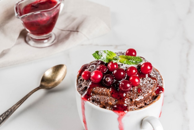 Winter, fall healthy breakfast or snack. The idea of a quick treat for Christmas. Chocolate mug cupkake in ceramic cup, with cranberry and sauce, powdered sugar. White marble table, copyspace