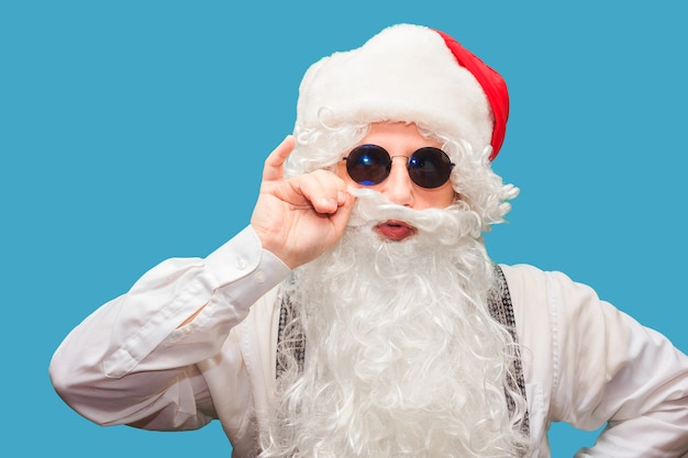 Winter fashion and sales. Portrait of a handsome man in a Santa hat, winking while standing against a red wall. The hipster is holding glasses and looking at the camera. Place for text.