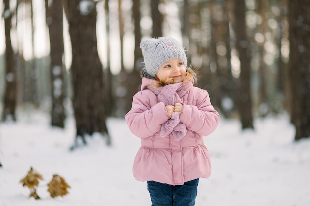 Inverno nella foresta. carina bambina sognante in un cappotto rosa in piedi nella foresta innevata con sfondo di alberi e guardando lontano. bambino sorridente che cammina in una gelida giornata invernale nella foresta