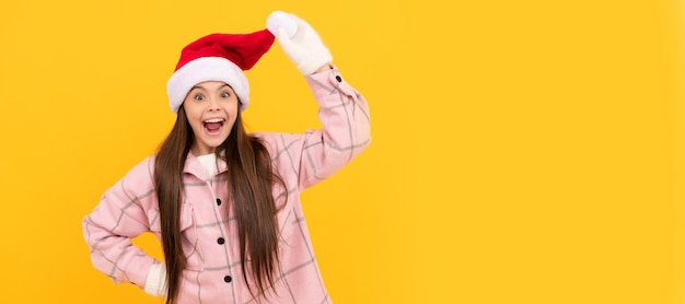 Winter girl amazed child in santa claus hat and winter mittens on yellow background Banner of christmas child girl studio kid winter portrait with copy space