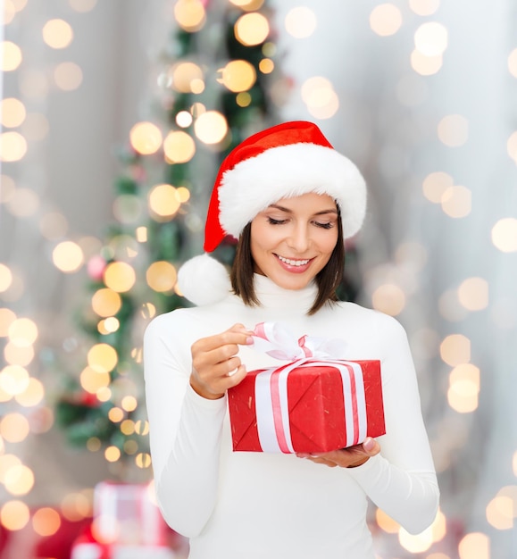 Inverno, felicità, vacanze e concetto di persone - donna sorridente in cappello di babbo natale con confezione regalo su sfondo soggiorno e albero di natale