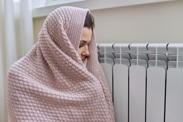 Зимний отопительный сезон Женщина в теплом свитере сидит в домашней комнате возле радиатора отопления