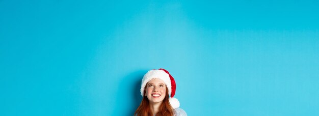Фото Зимние каникулы и канун рождества концептуальная голова милой рыжей девушки в новогодней шапке появляется снизу