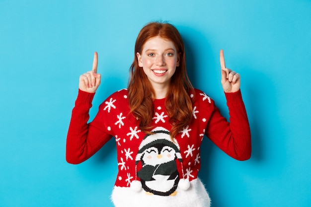 Winter holidays and celebration concept. Cute redhead girl in Christmas sweater, smiling and pointing fingers up at promo logo, standing over blue background