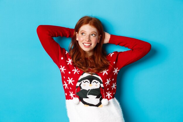 Winter holidays and celebration concept. relaxed teenage girl holding hands behind head and looking left at promo, resting on christmas eve, standing over blue background