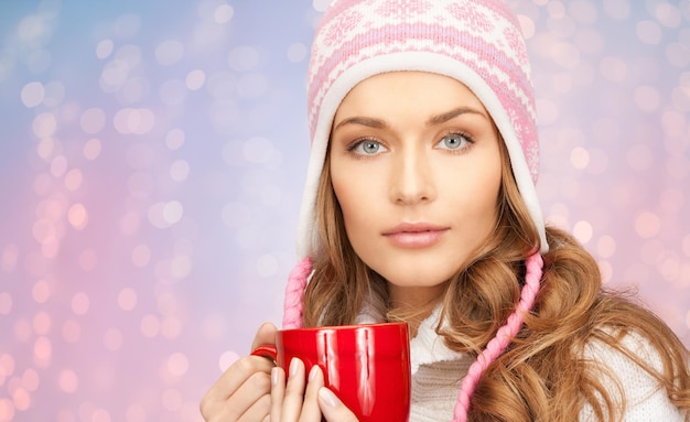 winter holidays, christmas, beverages and people concept - happy young woman in winter hat with red cup of tea over rose quartz and serenity lights background