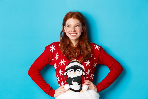 Photo winter holidays and christmas eve concept. beautiful teenage redhead girl in xmas sweater looking left at logo, smiling pleased, holding hands on waist, blue background