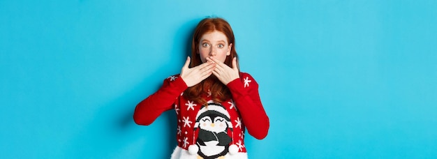 Winter holidays and christmas eve concept surprised redhead girl in cute sweater gasping and coverin