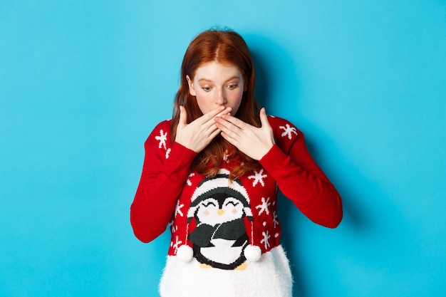 Зимние каникулы и концепция сочельника. Удивленная рыжая девушка, задыхаясь, с трепетом глядя вниз, глядя на логотип, стоит в рождественском свитере на синем фоне.