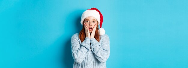 Foto vacanze invernali e concept della vigilia di natale ragazza rossa sorpresa con il cappello di babbo natale che fissa con la persona