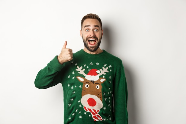 Зимние каникулы и рождество. Возбужденный кавказский парень показывает палец вверх и выглядит изумленным, стоя в зеленом свитере на белом фоне.