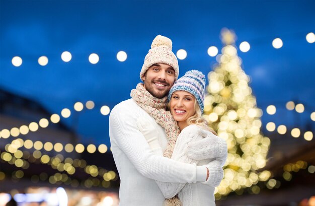 Winter, holidays, christmas and people concept - happy couple in hats and scarf hugging over night lights background