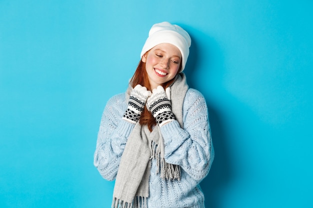 Концепция зимы и праздников. Симпатичная рыжая девушка краснеет и мечтательно смотрит в сторону, в теплой шапке, свитере и шарфе с перчатками, стоя на синем фоне.
