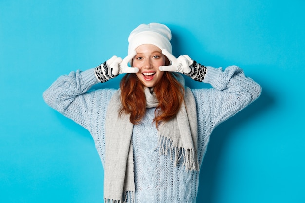 Концепция зимы и праздников. Симпатичная рыжая девочка-подросток в шапочке, перчатках и свитере показывает знак мира, смотрит налево в камеру и желает счастливого Рождества, стоя на синем фоне