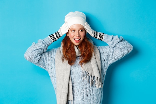 Winter and holidays concept. Happy redhead girl in beanie hat, scarf and gloves looking right and smiling, standing against blue background