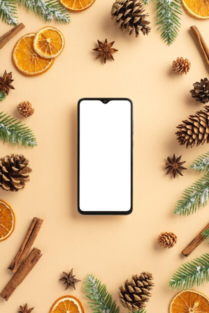 Photo winter holidays concept top view vertical photo of smartphone fir branches in frost dried orange slices pine cones anise and cinnamon sticks on isolated beige background with copyspace