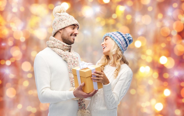 winter, holidays, couple, christmas and people concept - smiling man and woman in hats and scarf with gift box over lights background