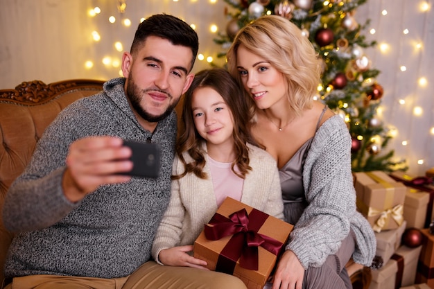 Winter holidays and family concept - happy young family taking selfie photo with smartphone in living room with decorated Christmas tree