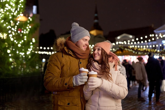 winter holidays, hot drinks and people concept - happy young couple with coffee at christmas market in evening