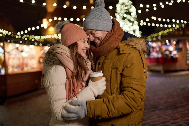 winter holidays, hot drinks and people concept - happy young couple with coffee at christmas market in evening