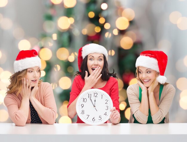 Winter, holidays, time and people concept - smiling women in santa helper hats with clock over christmas tree background