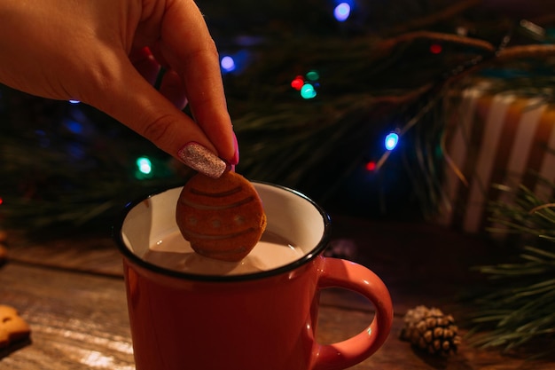 Winter holidays with latte and cookies Close up of unrecognizable woman dips delicious gingerbread cake into warm cup of coffee with milk on Christmassy pine and fairy lights background