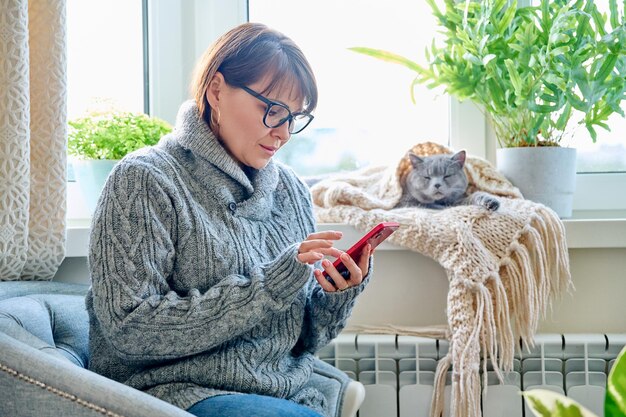 사진 겨울 가정 생활 방식 따뜻한 스웨터를 입은 중년 여성이 창문 근처의 좌석에 앉아 가열 라디에이터와 잠자는 고양이 창문 앞에 스카프를 입고 손에 스마트폰을 들고 휴식을 취하는 여성