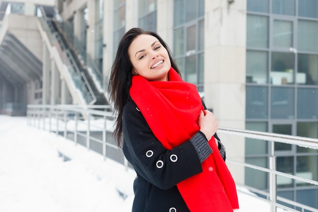 Winter, kerst, lifestyle, vakantie en mode concept - mooie jonge lachende vrouw in zwarte jas en rode sjaal poseren in winter stad. sneeuwen