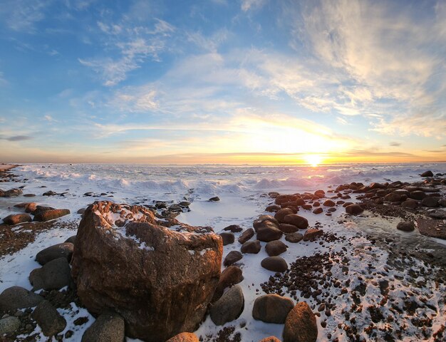 Winter landscape Panoramic view of beautiful sunset on bay List clouds over water in bright light Ice snow and rocks on coastline Setting sun is reflected in sea