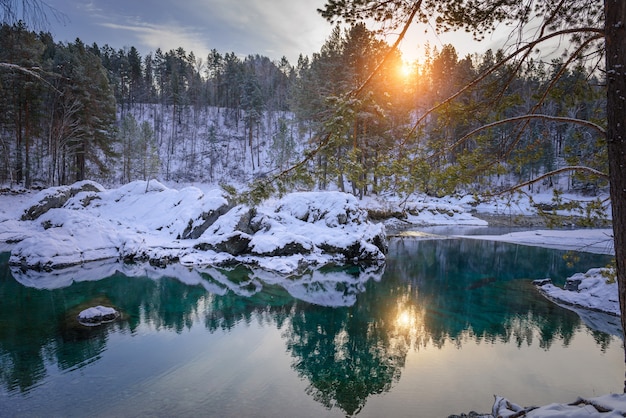 Winter landscape, small ice-free lake in the mountains among snow-covered forest. Trees are reflected in the lake water in evening sunlight