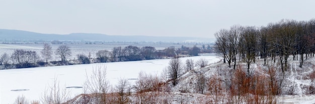 Winter landscape with snow-covered river and forest on a hill, panorama