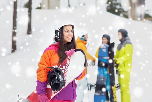 Winter, leisure, extreme sport, friendship and people concept -\
happy young woman in helmet with snowboard and group of\
friends