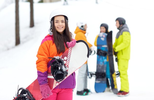 winter, leisure, extreme sport, friendship and people concept - happy young woman in helmet with snowboard and group of friends