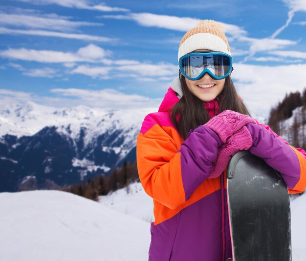 winter, leisure, sport and people concept - happy young woman in ski goggles with snowboard over snow and mountain background