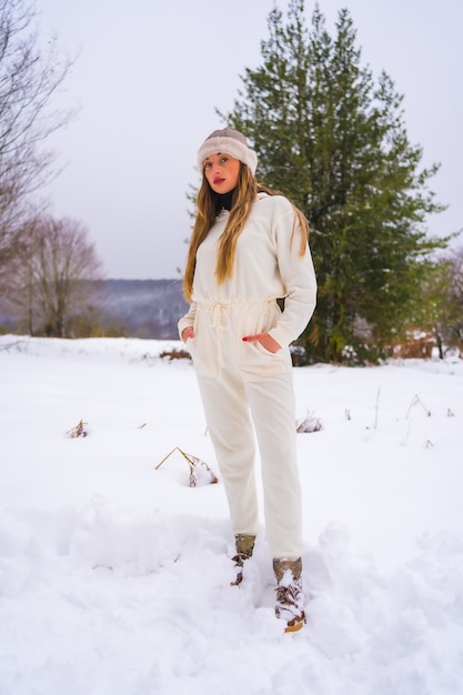 Winter levensstijl, portret van jonge mooie blonde blanke vrouw met witte winter outfit en wollen hoed in de sneeuw naast besneeuwde pijnbomen, vakantie in de natuur