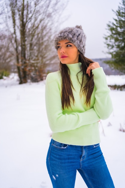 Winter lifestyle, portrait caucasian brunette with green outfit and woolen hat enjoying the snow next to snowy pine trees, vacation in nature