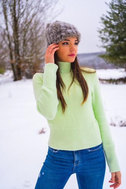 Winter lifestyle, portrait young brunette caucasian with green outfit and woolen hat enjoying the snow next to snowy pine trees, vacations in nature