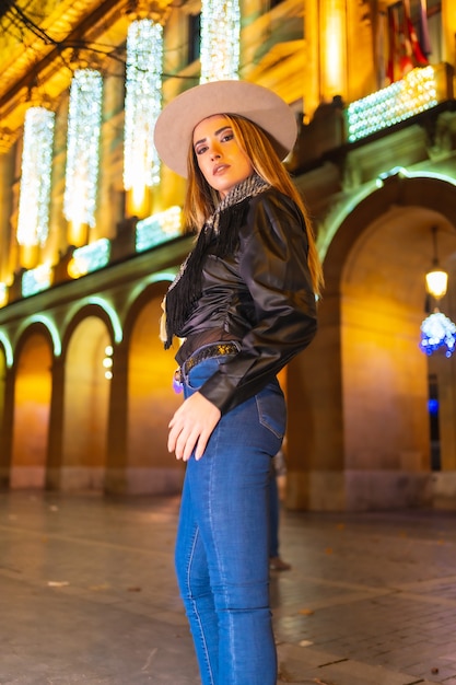 Winter lifestyle, young caucasian blonde with jeans, hat and a black jacket in the illuminated city at christmas