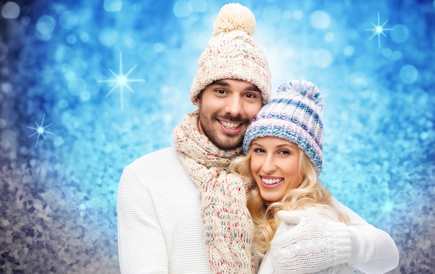 winter, mode, paar, kerst en mensen concept - lachende man en vrouw in hoeden en sjaal knuffelen over blauwe glitter en vakantie lichten achtergrond