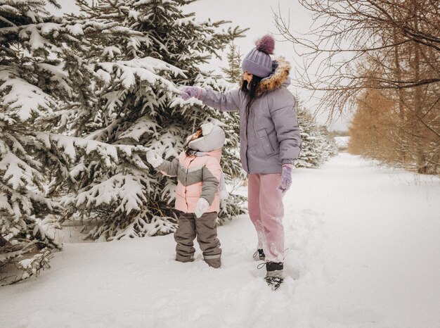 Winter outdoor recreation. Happy mother shakes off snow from a branch on a child standing among snow-covered fir trees